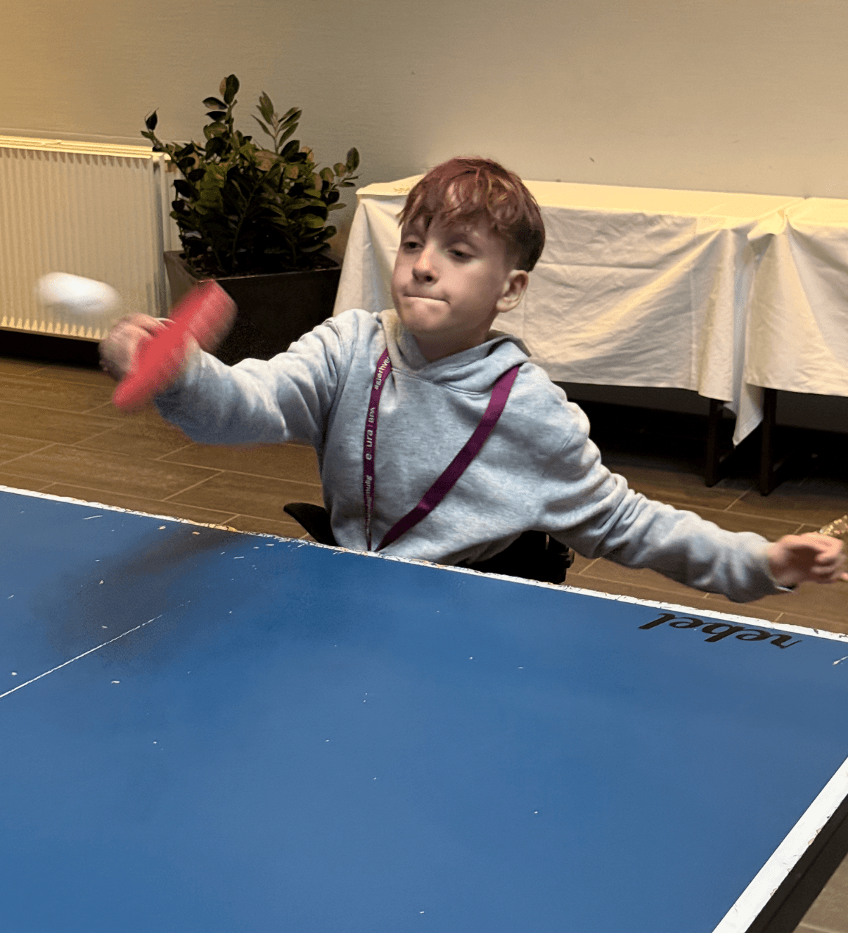 Arvid spiller bordtennis med fokus og energi i et innendørs fellesrom, iført grå hettegenser og lanyard – et uttrykk for selvstendighet og aktivitet.