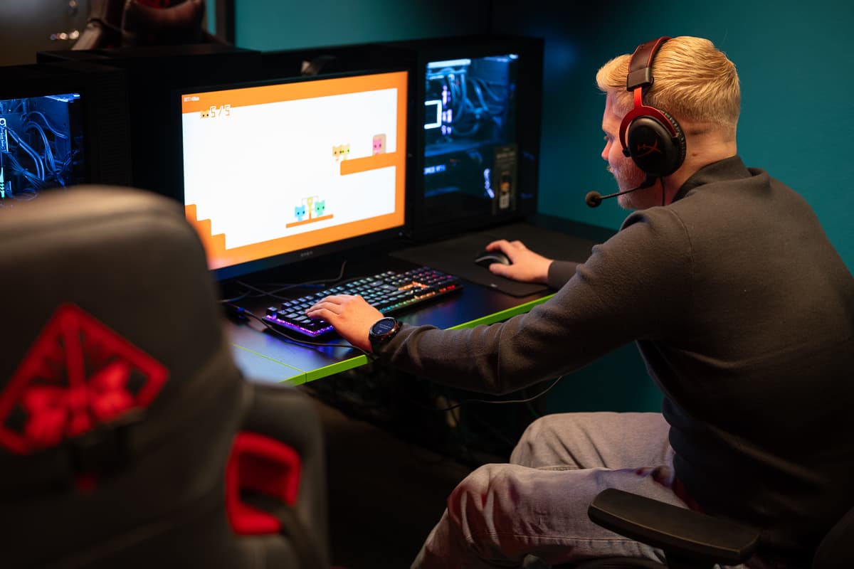 Person med headset spiller fargerikt dataspill med tegneseriefigurer på skjerm. Gamingstasjonen har RGB-tastatur, kraftig PC og stol med OMEN-logo. Spillet viser "5/5" øverst.