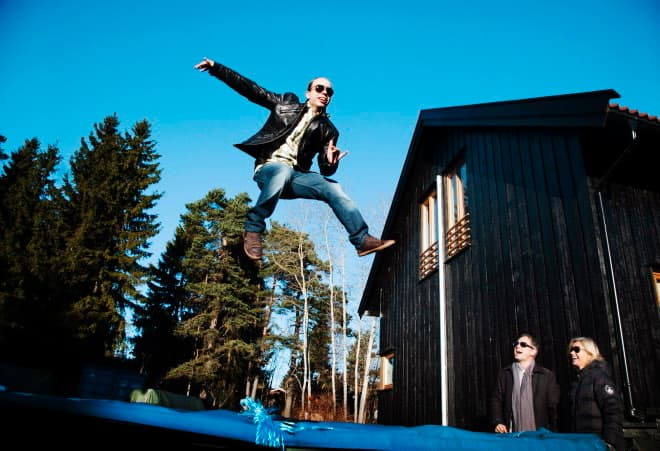 Anders i svart skinnjakke og jeans hopper på trampoline i hagen, med hus og furutrær i bakgrunnen.