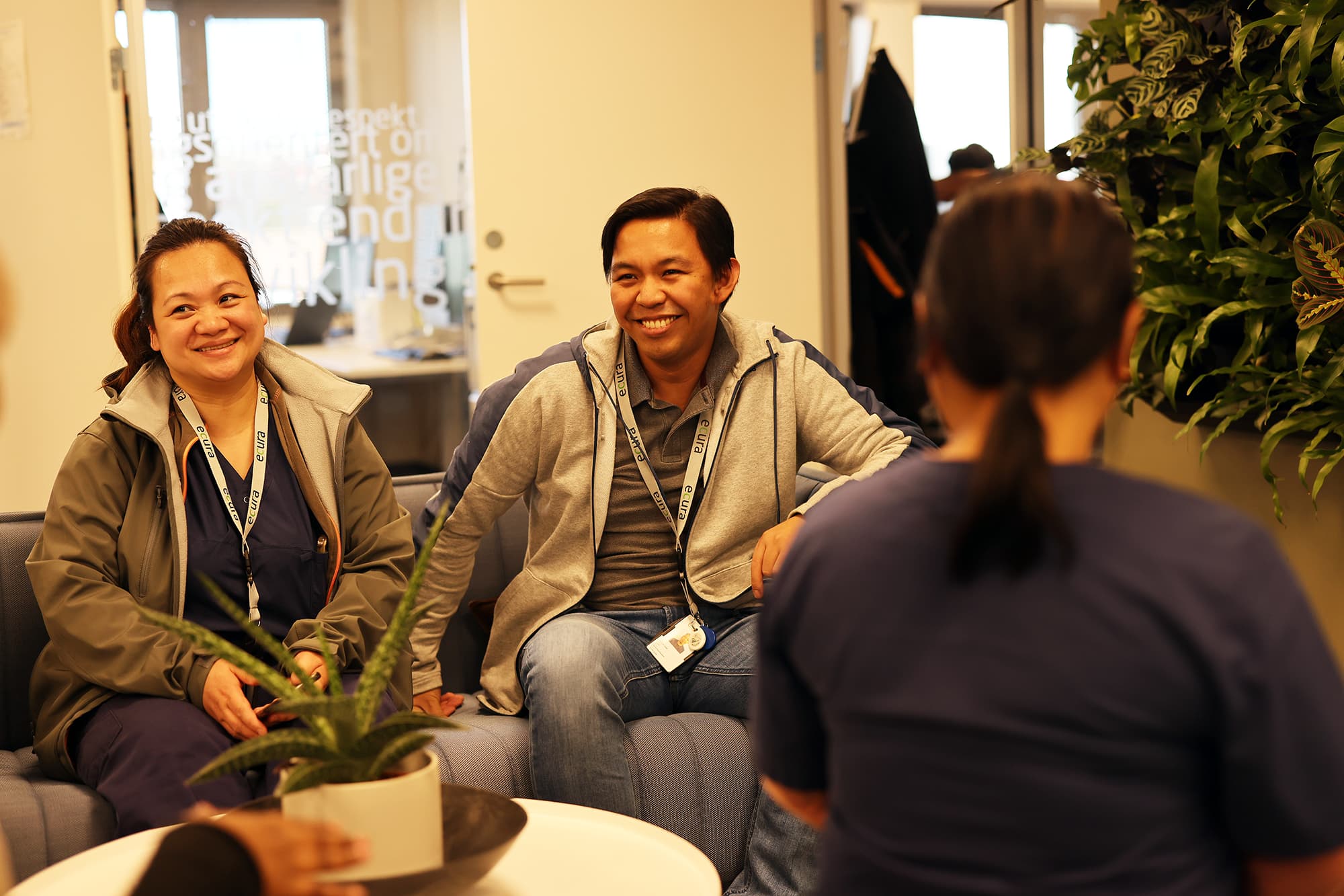 Fire personer i samtale i et lyst og uformelt kontorlandskap, med grønn plantevegg, glassdør med tekst og bord med potteplante i forgrunnen. To personer med navneskilt sitter på sofa.