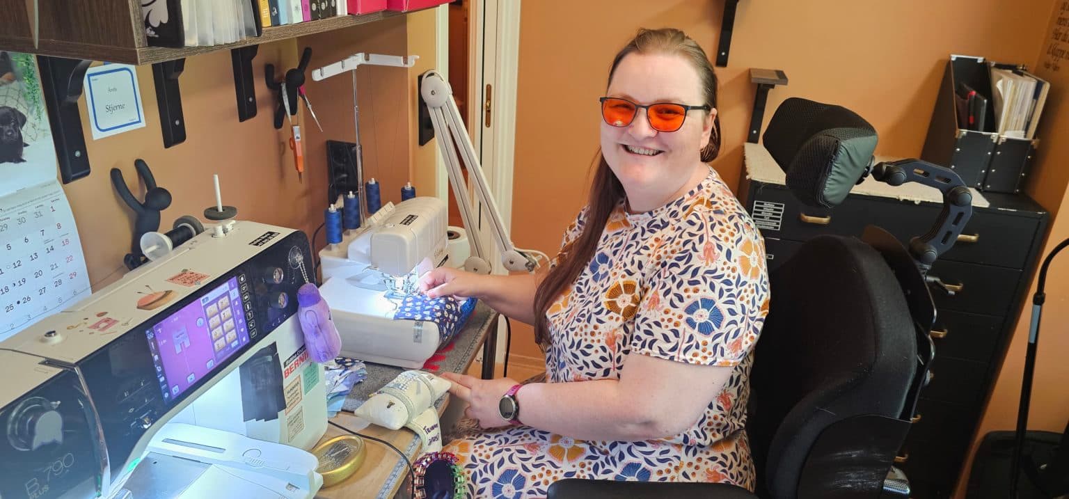 Halldis Marie sitter i rullestol smiler mens hun syr blåmønstret stoff ved en moderne symaskin i et ryddig og personlig syrom. Hen har oransje briller og blomstrete kjole. Rommet er utstyrt med verktøy, trådspoler, bøker og en veggkalender for juni. Navnet "Karla Stjerne" vises på veggen.