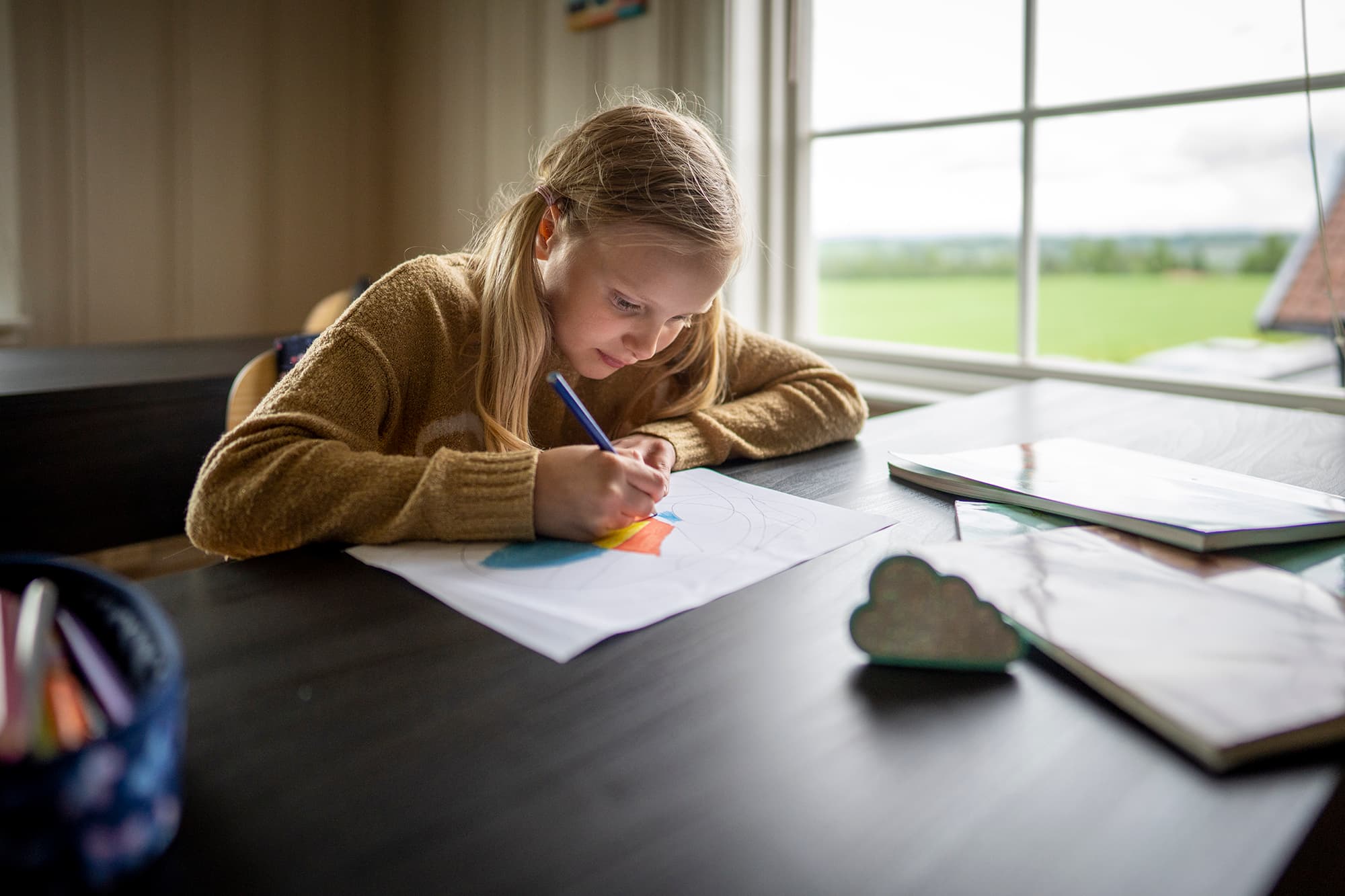 Barn med lyst hår fargelegger tegning med fargeblyanter ved bord ved stort vindu. Utsikt mot grønn eng og overskyet himmel. Bøker, blyanter og skyformet pynt på bordet.