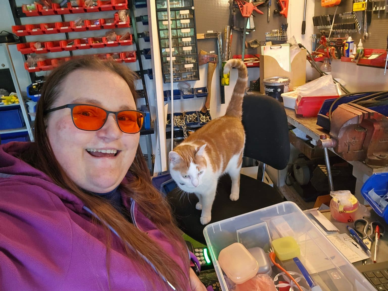 Halldis Marie med oransje briller og lilla hettegenser smiler i et verksted, mens en hvit og oransje katt står på en stol ved siden av. Bakgrunnen viser ryddige verktøy, bokser og hyller. På bordet foran dem ligger en plastboks med pensler og småting.