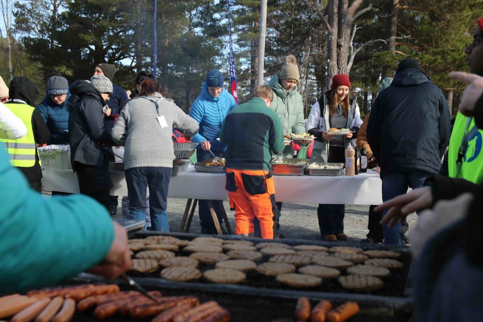 Folk samles rundt langbord med hvit duk og matservering i skogsområde. I forgrunnen grilles hamburgere og pølser. Deltakerne har på varme klær og hjelper hverandre med mat.