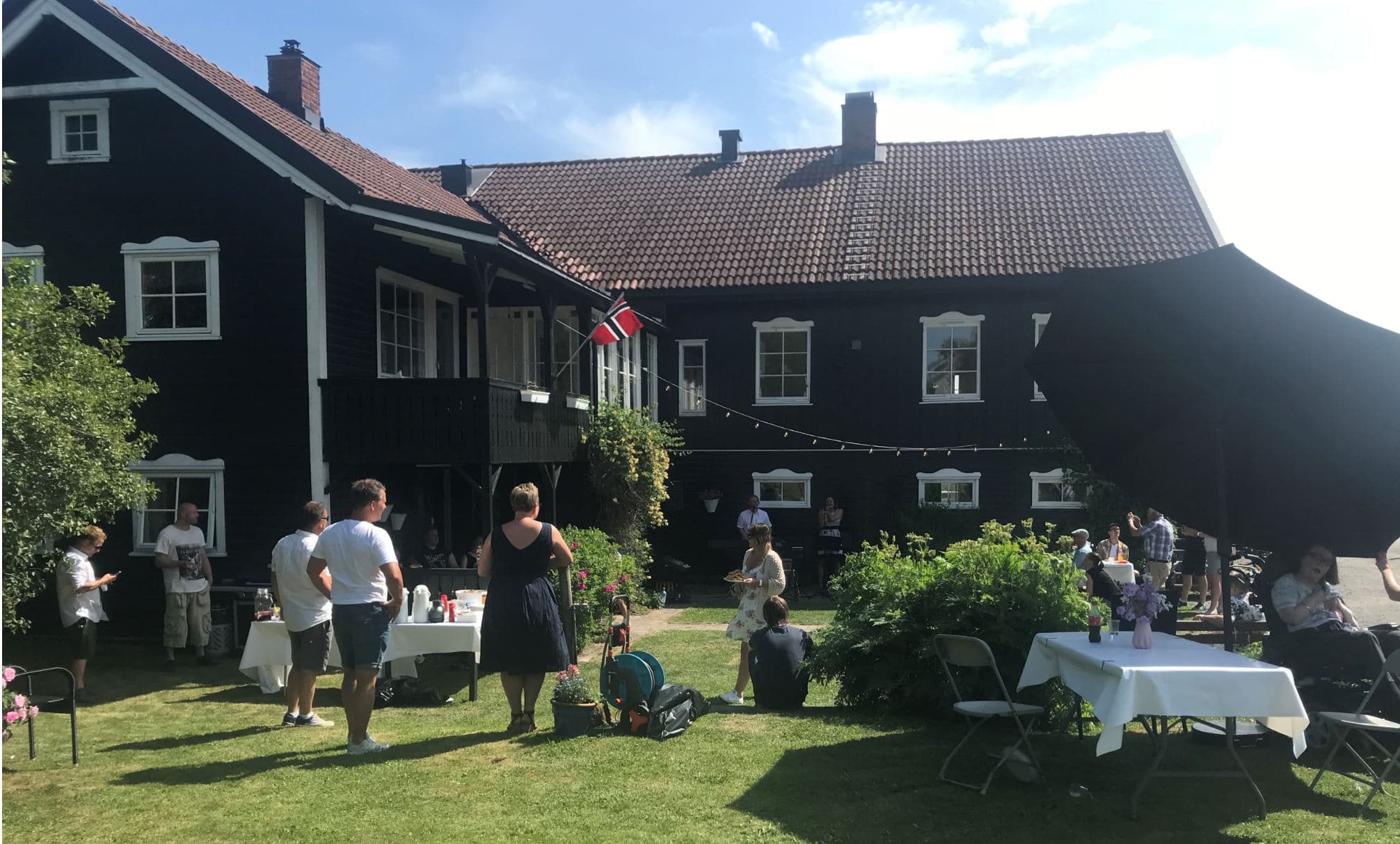Folk samles i hagen foran et stort, mørkt trehus med hvite detaljer og rød takstein. Norske flagg og hvite duker på bordene tyder på fest eller sosial markering. Lyslenker henger over plassen, og gjestene står og sitter i små grupper mens de prater og koser seg. Solskinn, blomster og grønt gir en livlig og hyggelig stemning.