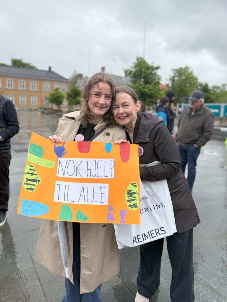 To personer står ute i regnet og holder en oransje plakat med teksten "NOK HJELP TIL ALLE" og fargerike tegninger. Bakgrunnen viser våt asfalt, folk og eldre bygninger.