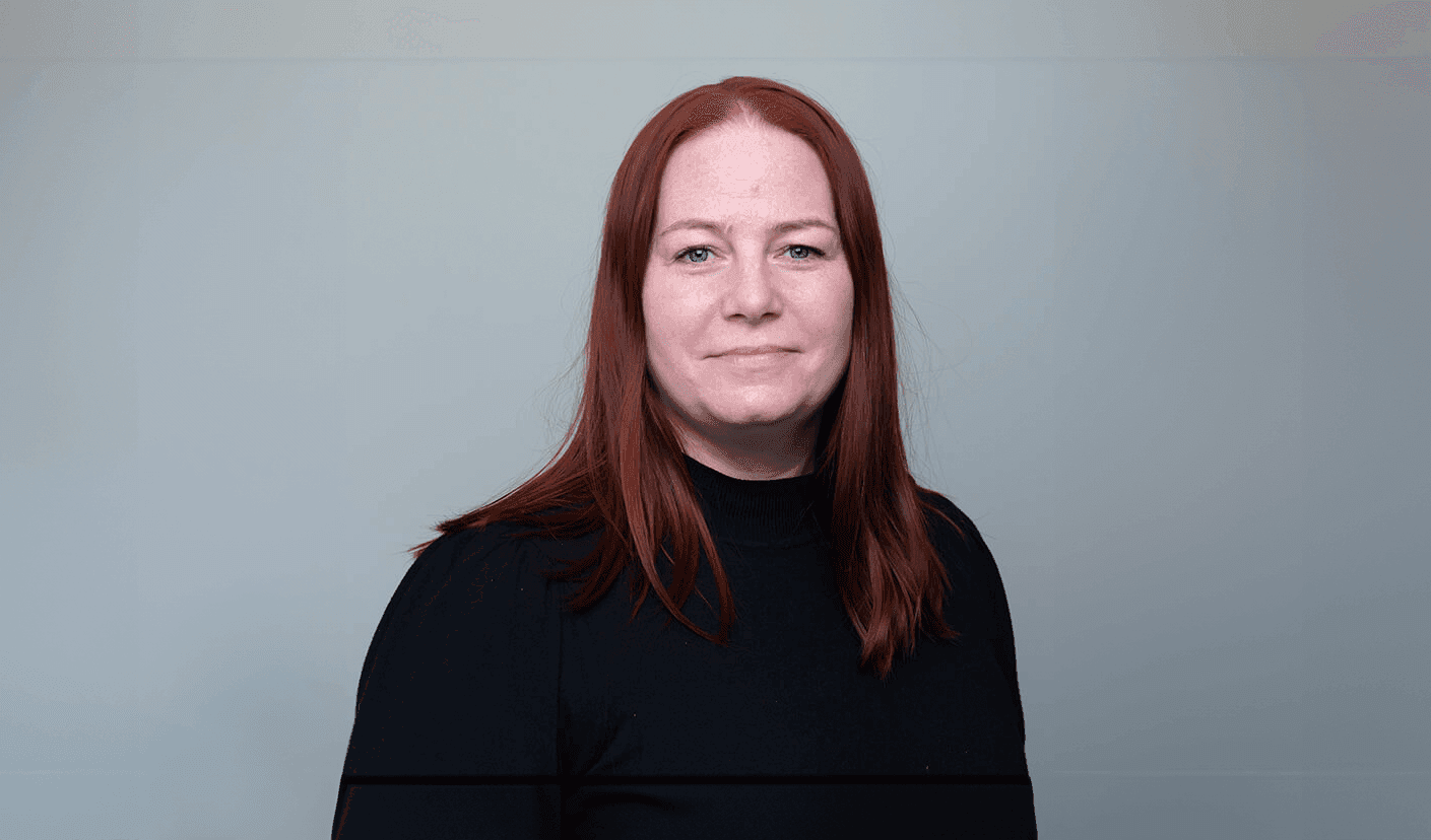 Person med langt rødt hår og svart topp står foran lys grå bakgrunn og ser rett mot kamera. Bildet har nøytral bakgrunn og fremstår som et profesjonelt portrett eller profilbilde.