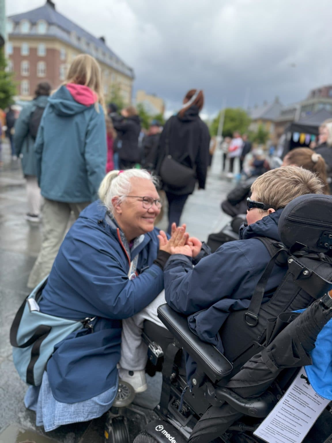 Kvinne med hvitt hår og briller kneler og holder hender med en person i elektrisk rullestol. Begge smiler. Bak dem sees folk, klassiske bygninger og overskyet himmel.