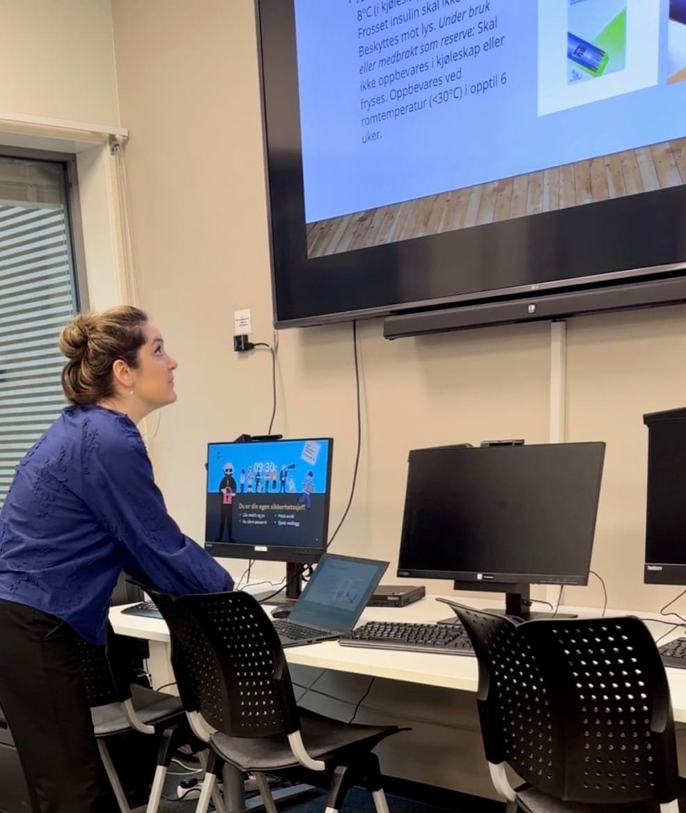 Kvinne står foran skjerm med bilder av insulinpenner og norsk tekst om oppbevaring. Dataskjermer og undervisningsmiljø i bakgrunnen.