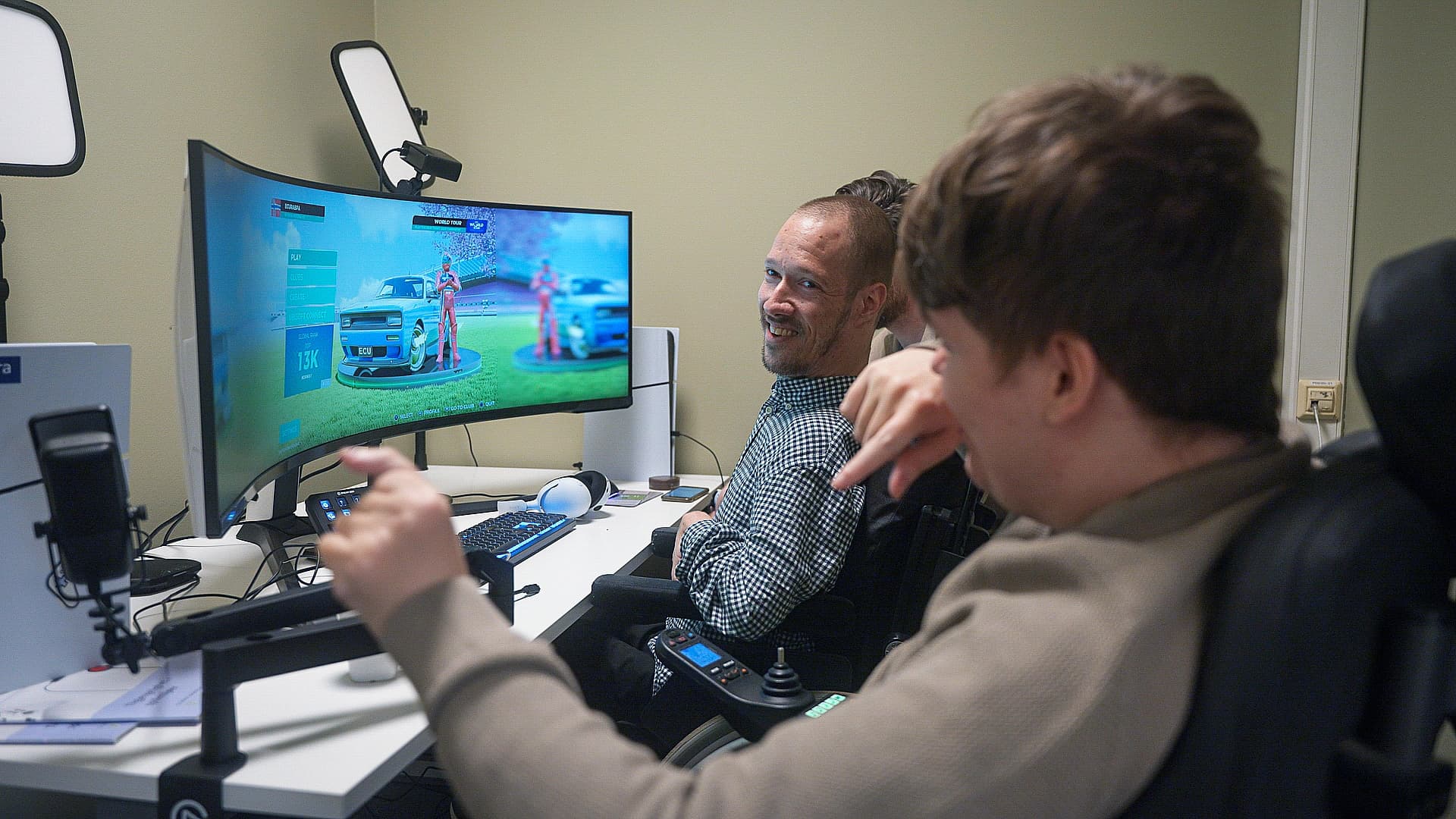 To personer spiller bilspill på stor buet skjerm. Én peker mot skjermen, den andre smiler og ser bakover. En av dem bruker rullestol og adaptive kontroller. Pulten har tastatur, mikrofon og lysutstyr.