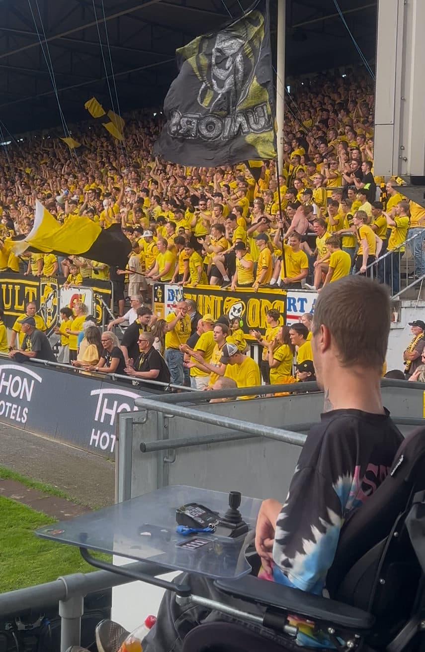 Stadion fylt med gule og svarte supportere, flagg og bannere, med en person i elektrisk rullestol i forgrunnen – et bilde på inkluderende idrettsglede og fellesskap.