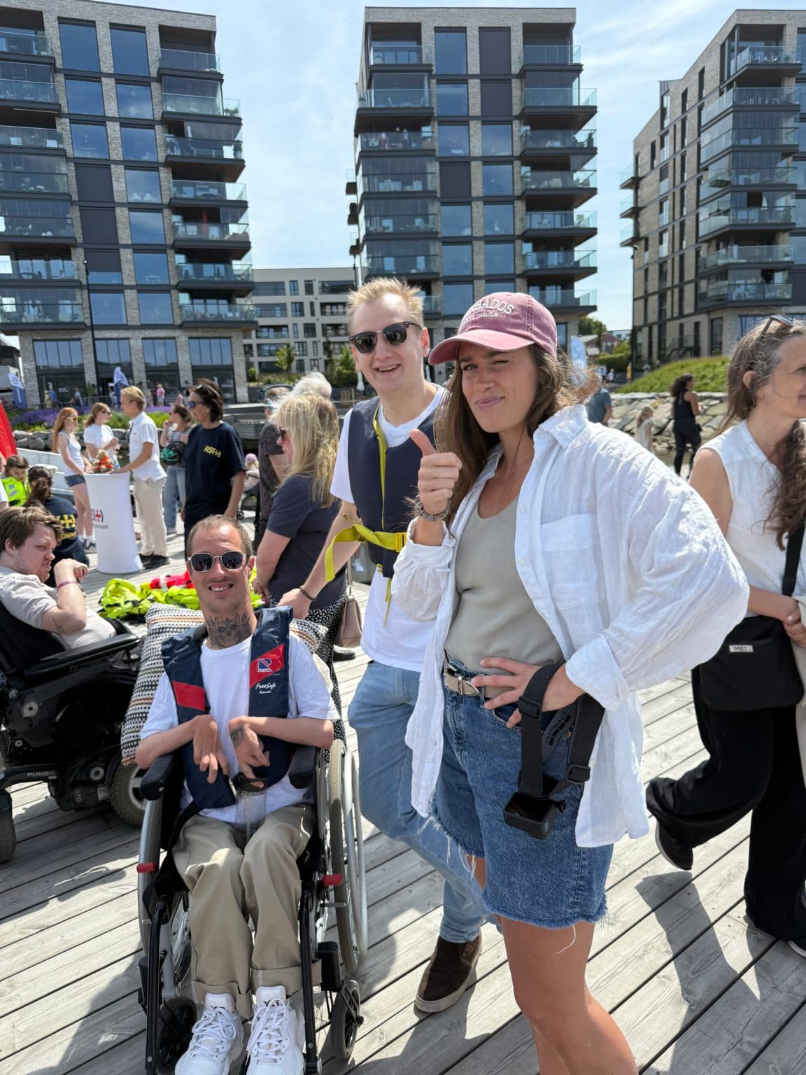 Tre personer smiler og poserer på en treplatting foran moderne leilighetsbygg. En person i rullestol har på hvit skjorte og beige bukser, en annen står bak med solbriller og gul stropp, og den tredje gir tommel opp med caps og denimshorts.