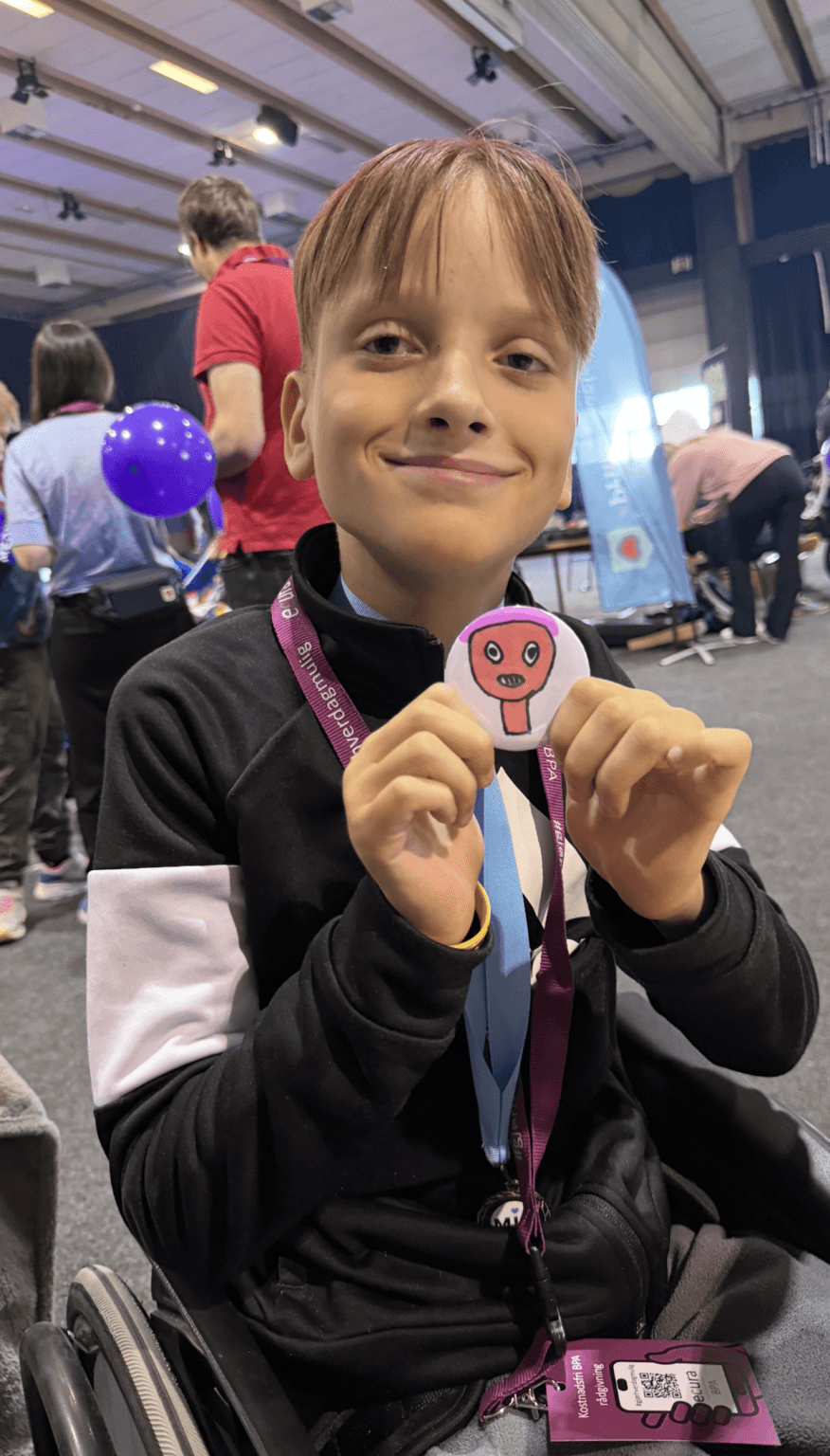 Arvid smiler og viser frem en kreativ button under Elevredagen, omgitt av deltakere og ballonger – et bilde på engasjement og inkludering.