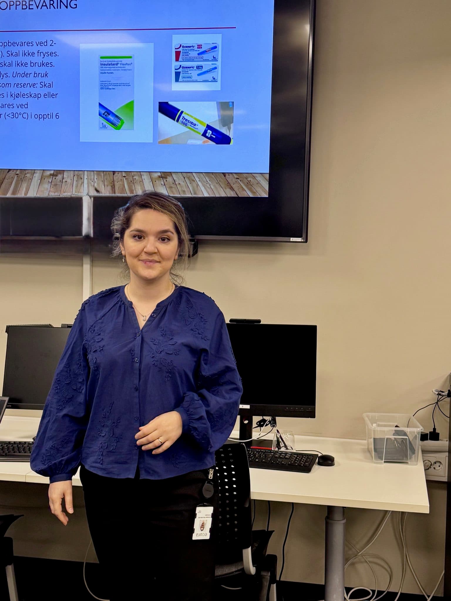Kvinne i blå bluse står ved pult med dataskjermer og ser mot stor skjerm med insulin-instruksjoner. Moderne undervisningsrom med stoler, lydpanel og presentasjon på norsk.