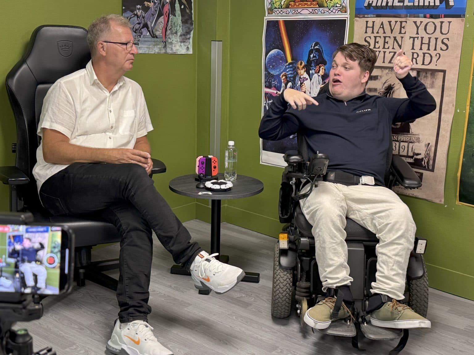 To personer i samtale i et gamingrom, én i gamingstol og én i elektrisk rullestol. Mellom dem står et bord med Nintendo Switch og gamingutstyr, omgitt av plakater fra Star Wars og Harry Potter – et inkluderende møte med fokus på spillkultur og fellesskap.