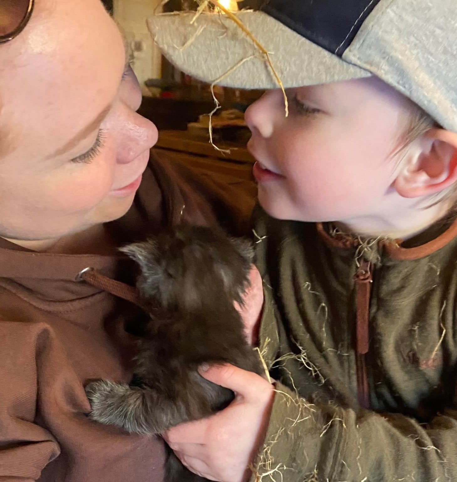 Mariella og Lasse deler et varmt øyeblikk mens de holder en liten svart kattunge. Mariella har brun hettegenser, Lasse har grønn jakke med strå og grå caps. De er tett sammen i et rustikt miljø.