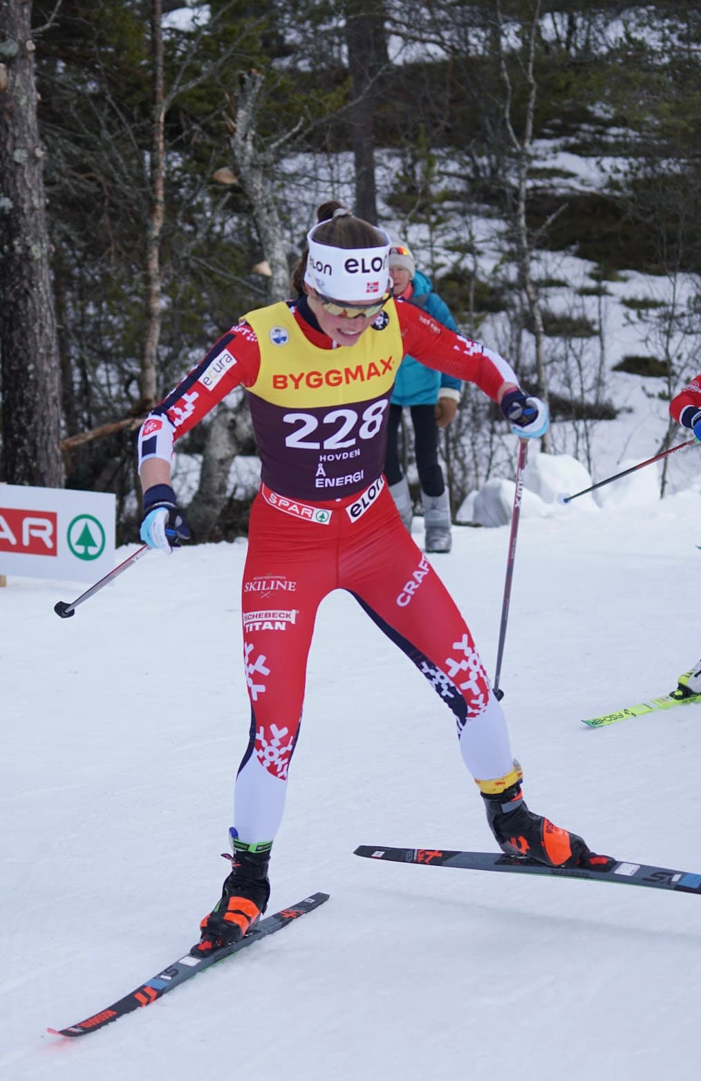 Hedda Bakkemo med startnummer 228 i rød og hvit drakt under skirenn, omgitt av snø, trær og tilskuere.