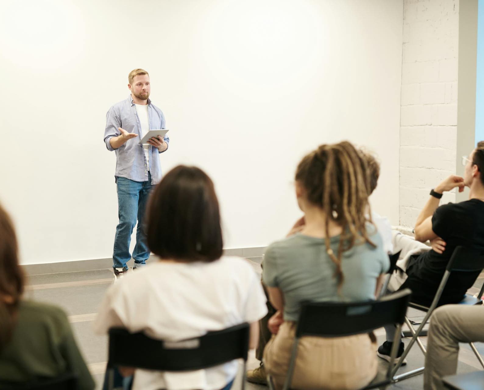 Mann i lyseblå skjorte holder presentasjon med nettbrett foran et lyttende publikum i et lyst, minimalistisk rom.