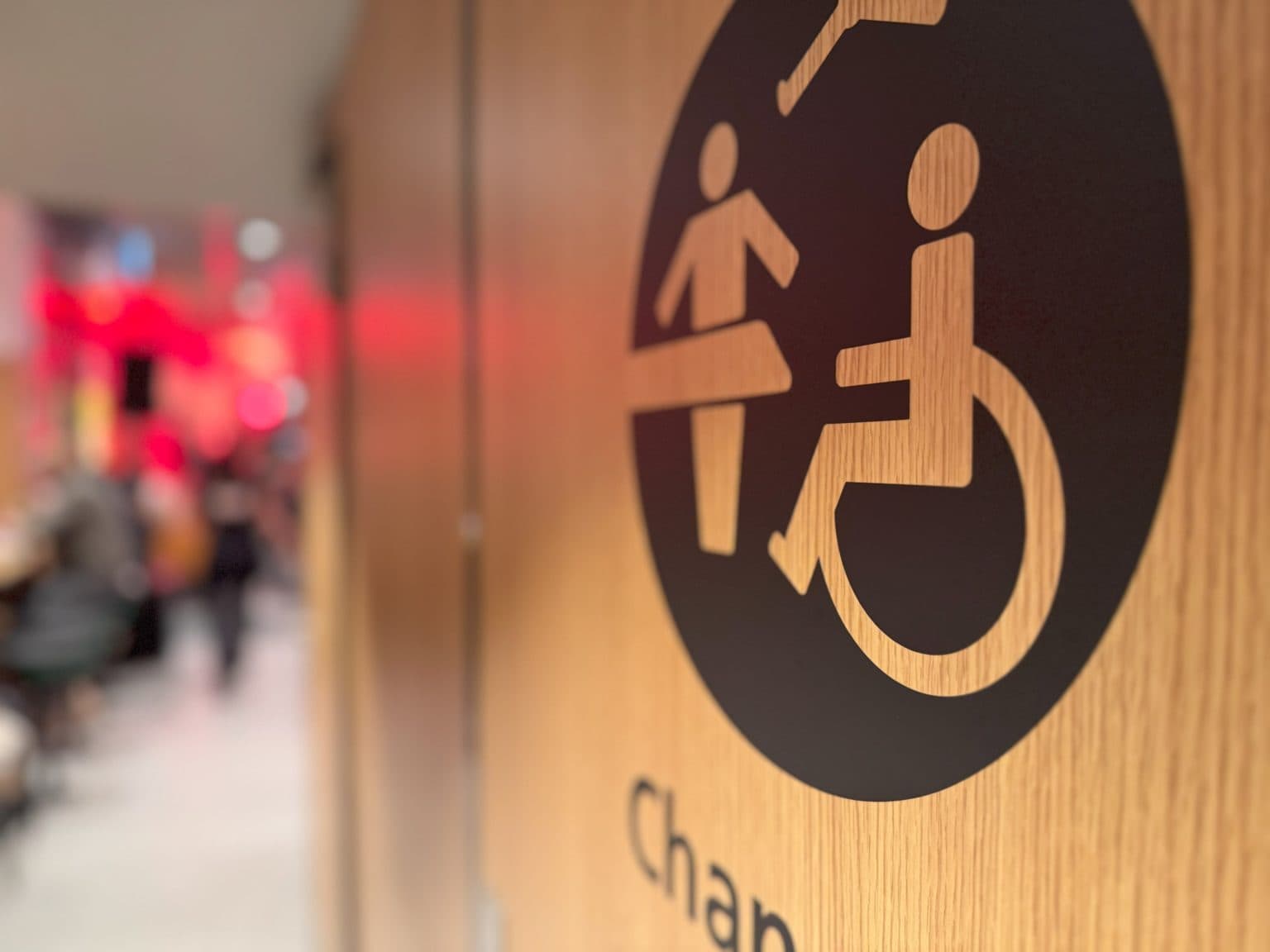 Nærbilde av en tredør med svart skilt som viser symboler for rullestolbruker, stående person og person på stellebord. Delvis tekst "Chan" vises under. I bakgrunnen skimtes en offentlig korridor med folk og rød belysning.
