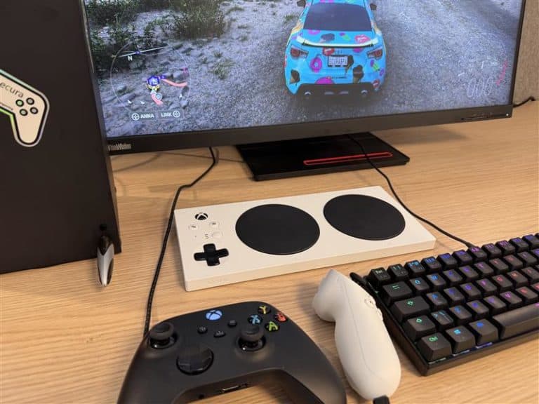 Gamingoppsett med skjerm som viser bilspill, Xbox Adaptive Controller, vanlig kontroller og RGB-tastatur. På skjermen vises karakterene "ANNA" og "LINK". En PC med Ecura-klistremerke står ved siden av.