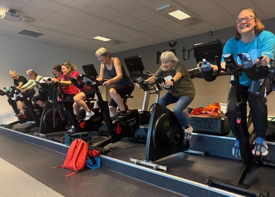 Syv personer trener på stasjonære sykler i et treningssenter. De sitter på rekke på en opphøyd plattform, med digitale skjermer og vannflasker på syklene. Deltakerne har på treningsklær og virker engasjerte. Bak dem vises vegg med teksten "trening".