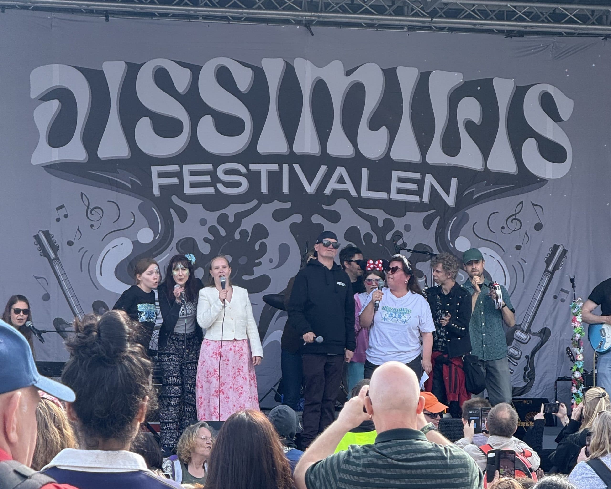 Gruppe mennesker opptrer eller snakker på scenen under Dissimilis Festivalen. Bak dem vises fargerik bakgrunn med gitarer, noter og festivalnavnet. Publikum i forgrunnen tar bilder og følger med.