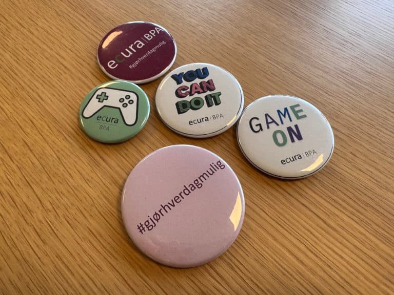 Fem runde buttons ligger på en treflate, hver med fargerik design og motiverende budskap knyttet til Ecura BPA. Tekster inkluderer "YOU CAN DO IT", "GAME ON", "#gjørhverdagenmulig" og grafikk som spillkontroller.