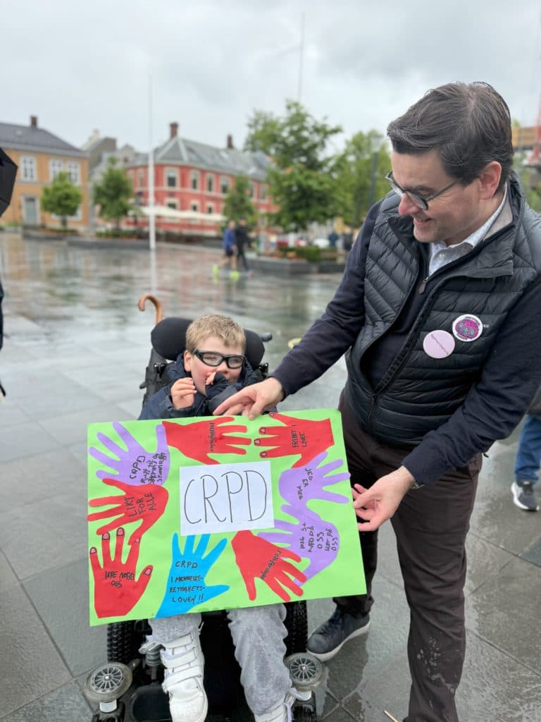 Barn i rullestol og voksen holder en fargerik plakat med teksten "CRPD" og utklipte hender i rødt, lilla og blått med budskap som "INDEPENDENCE", "RIGHTS" og "JEG HAR RETTIGHETER OG OSS". De står på våt brostein i en offentlig plass med bygninger og trær i bakgrunnen..