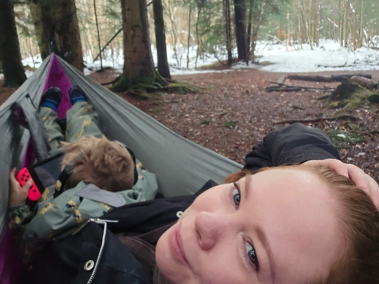 Mariella og Lasse ligger i hengekøye i skogen. Den ene ser mot kamera, den andre holder en rød Nintendo Switch-kontroller og har på hodetelefoner. Bakken er dekket av løv og snø, og høye trær omgir dem.