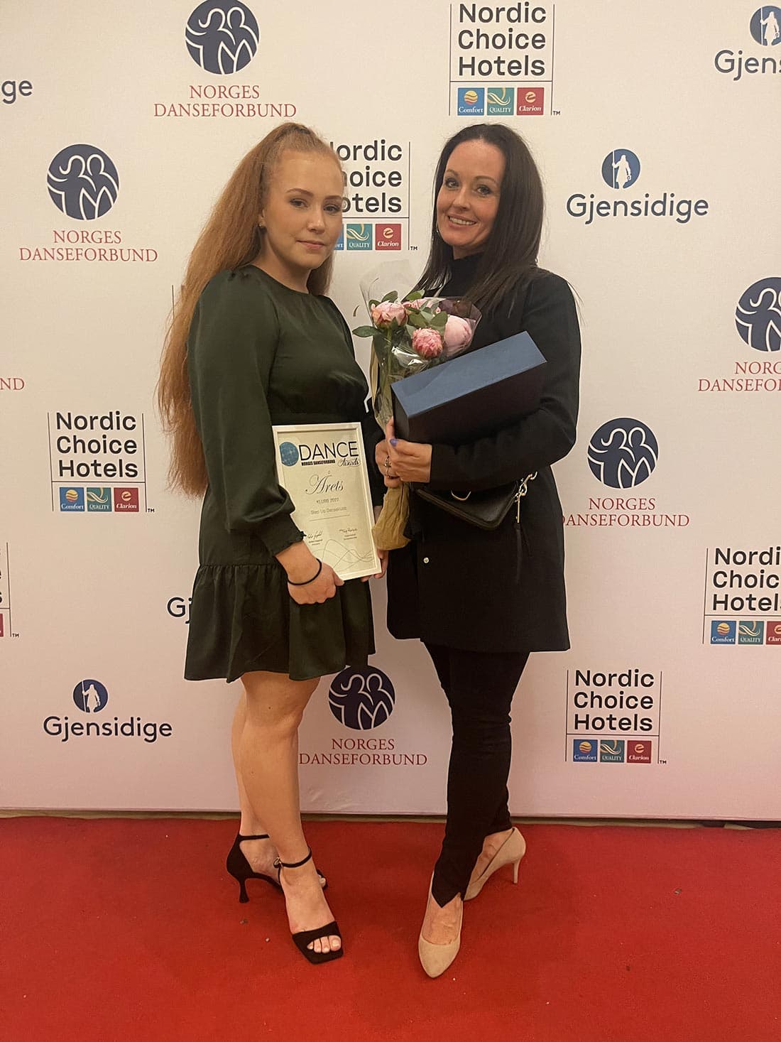 To personer står på rød løper foran backdrop med logoer fra "Norges Danseforbund", "Nordic Choice Hotels" og "Gjensidige". Den ene holder diplom med teksten "DANCE Awards" og blomster, den andre holder gaveeske og bukett.