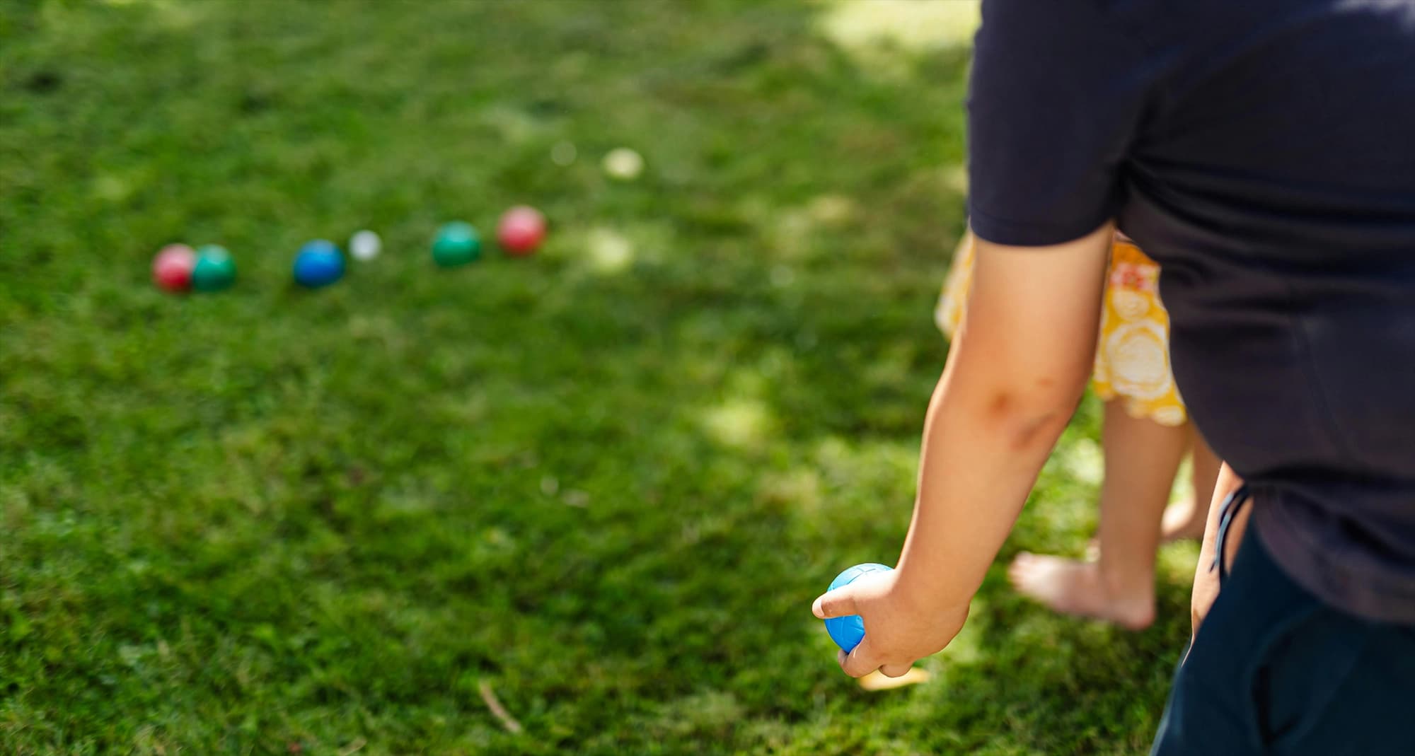 Person holder en blå boccia-ball på en gressplen, klar til å kaste. Flere fargede baller og en liten hvit pallino ligger spredt i bakgrunnen. En annen person står barbeint ved siden av. Uformell utendørs aktivitet i sollys.