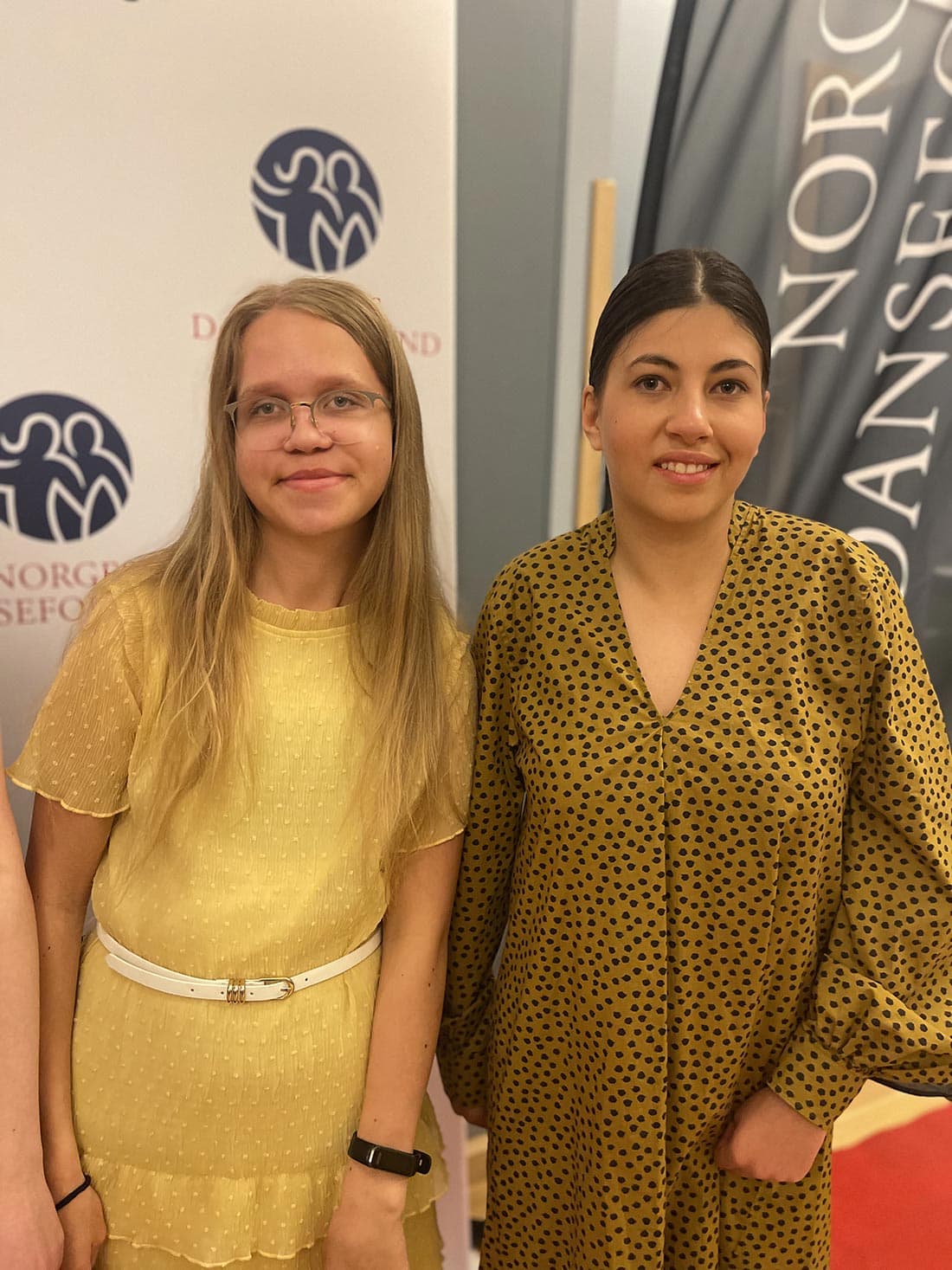 To personer står side om side foran backdrop og banner med logo og teksten "NORGES DANSEFORBUND". Den ene har lysegul kjole og briller, den andre har sennepsgul kjole med svarte prikker.