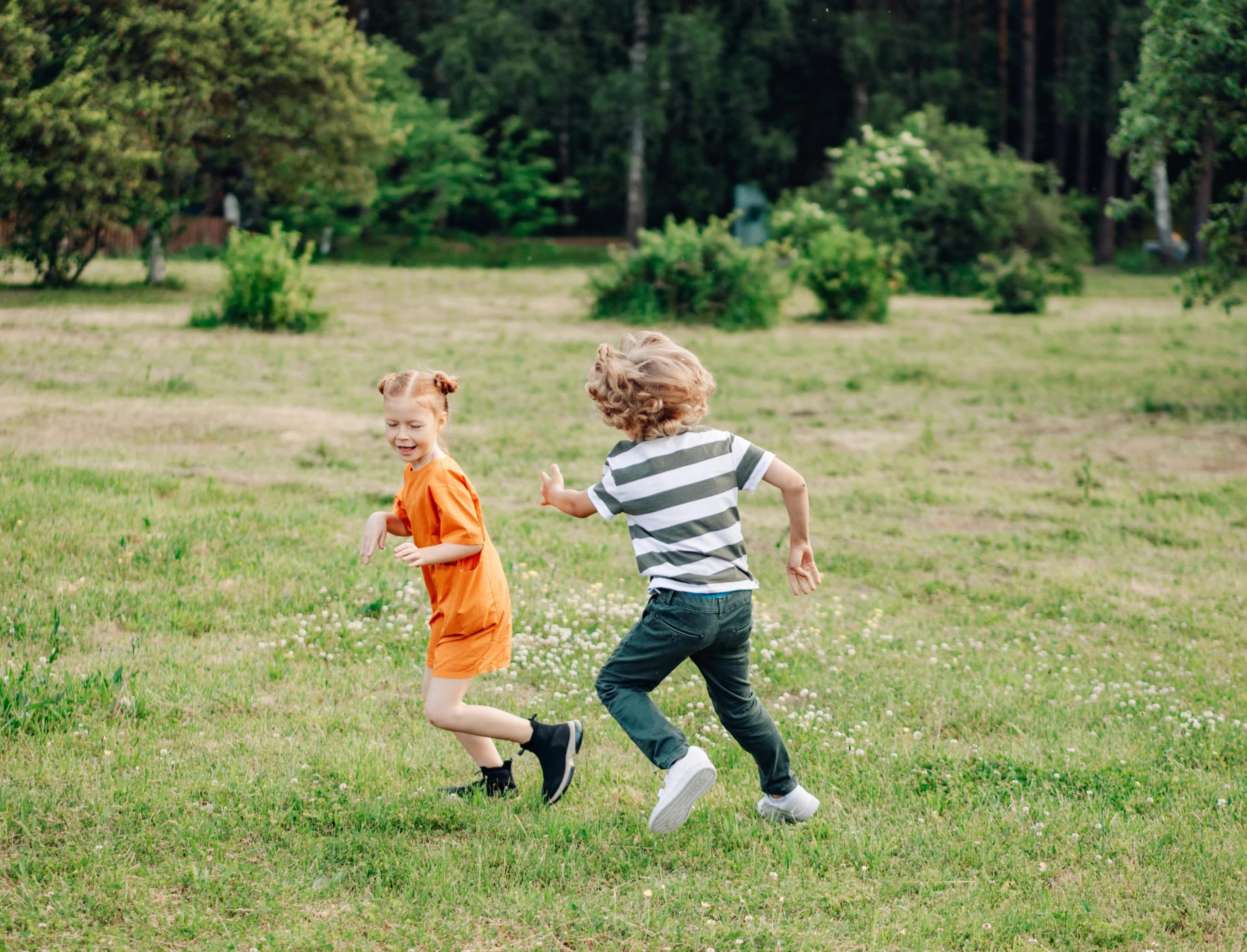 To barn, én i oransje antrekk og én i stripete skjorte, løper og leker på en gresslette omgitt av busker og trær i en frodig park.