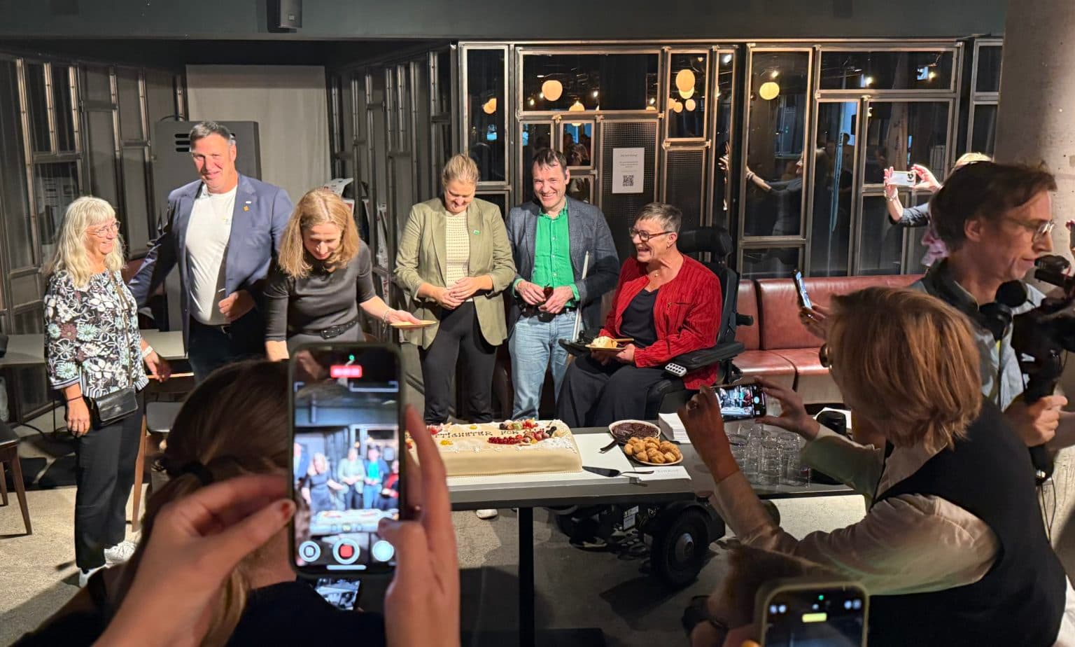 Gruppe mennesker samlet rundt en stor kake, der en person i elektrisk rullestol holder et kakestykke – et bilde på feiring, inkludering og samhold.