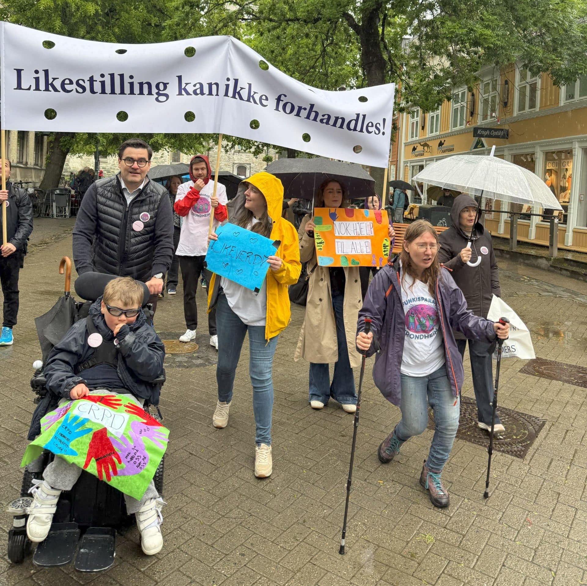 Mangfoldig gruppe mennesker deltar i en demonstrasjon med bannere og plakater. En person i rullestol og andre med hjelpemidler bærer budskap som "Likestilling kan ikke forhandles!" og "NOK HJELP TIL ALLE".