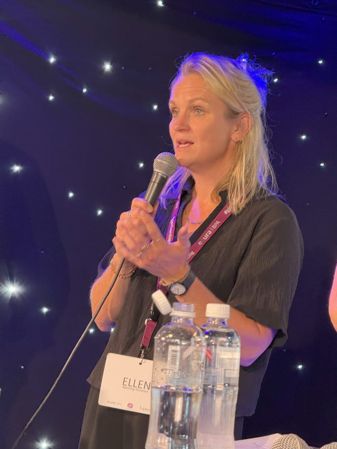 Kvinne med mikrofon og navneskiltet «Ellen Rønning-Arnesen» taler under konferanse, omgitt av stjernelys og vannflasker – et bilde på personlig engasjement og offentlig formidling.