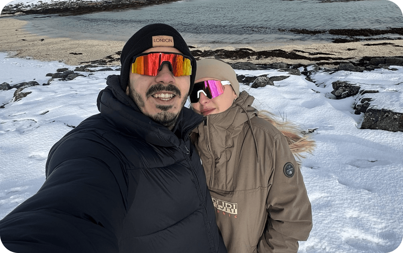 Selfie av to personer i vinterklær og reflekterende solbriller, med snø og vann i bakgrunnen.