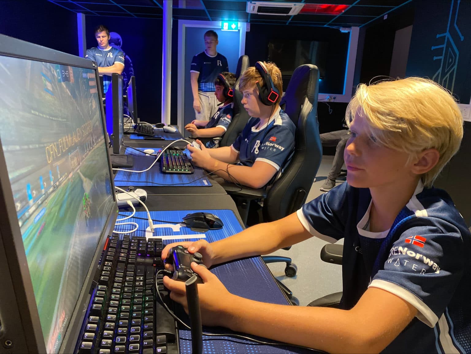 Ungdommer i matchende drakter spiller "Rocket League" i profesjonelt e-sportmiljø. De sitter ved gamingstasjoner med skjermer, tastatur, kontroller og headset. Draktene har logoer og dansk flagg. Blå belysning og ergonomiske stoler gir konkurransepreg.
