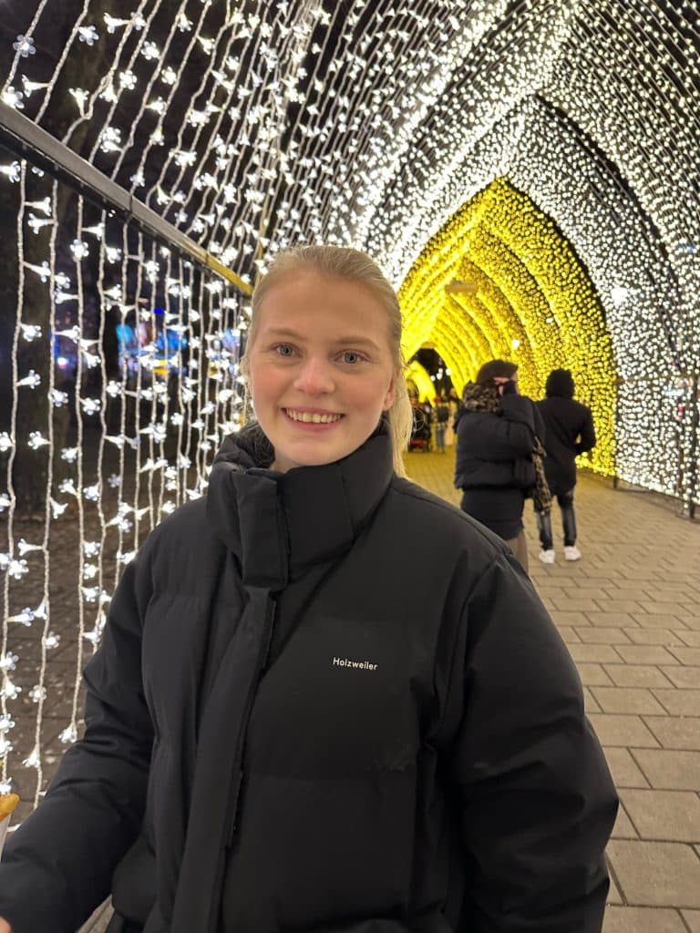 Person med svart vinterjakke fra Holzweiler står foran en tunnel av små hvite og gule lys i buemønster. Bak sees folk som går gjennom lysinstallasjonen på en brolagt sti.
