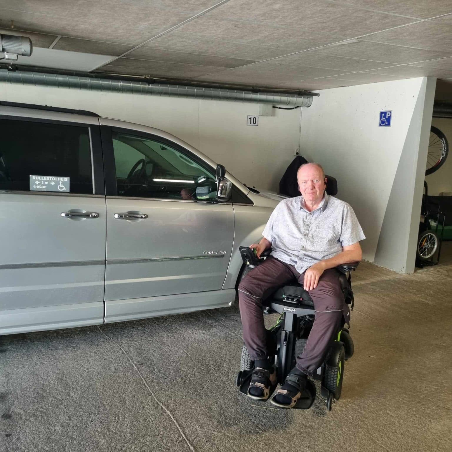 Mann i motorisert rullestol ved Chrysler-minivan med rullestolheis. Parkeringsplass med blå HC-skilt og nummer 10.