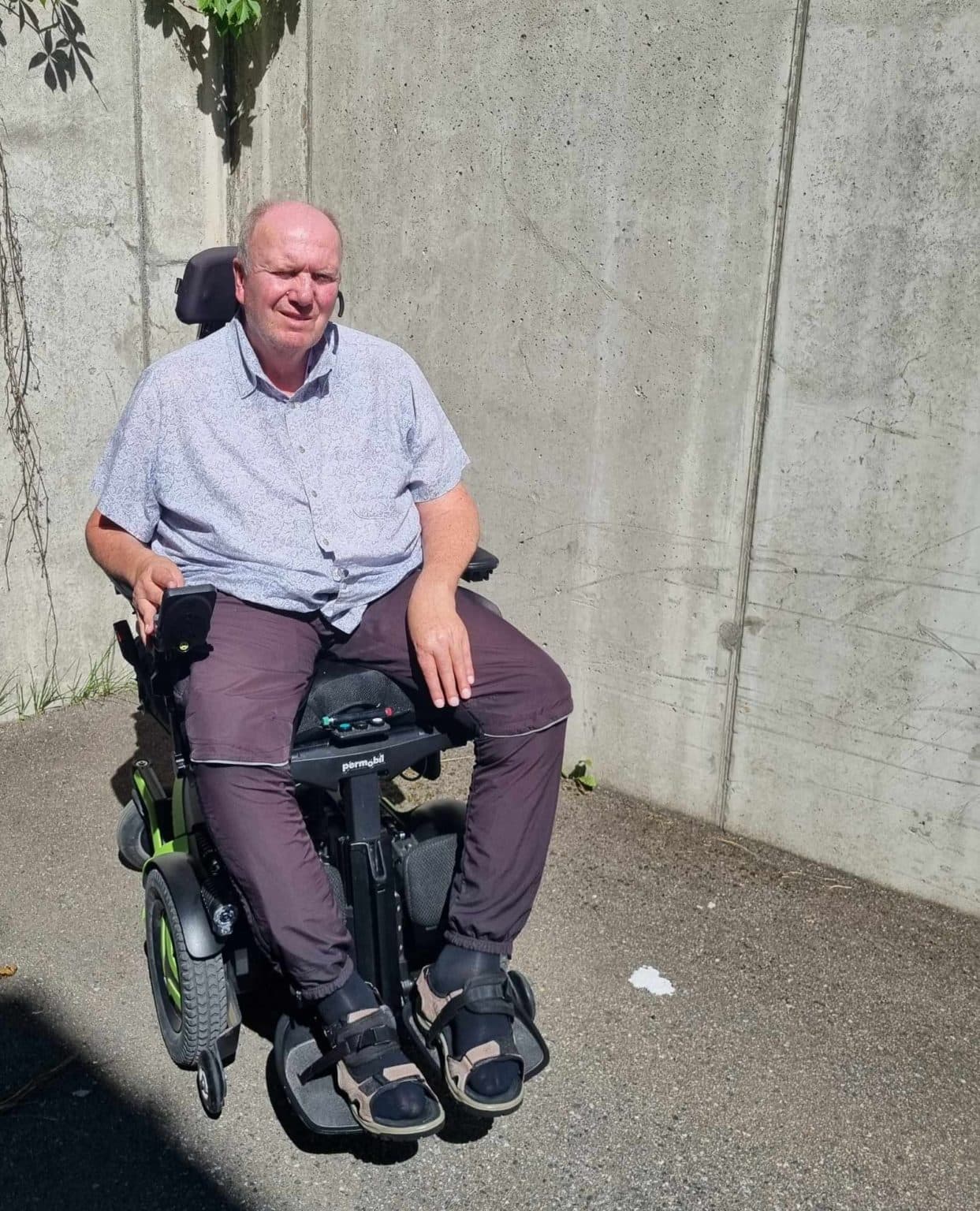 Person i motorisert rullestol på asfalt ved murvegg med slyngplanter. Lys skjorte, mørke bukser og sandaler med sokker.