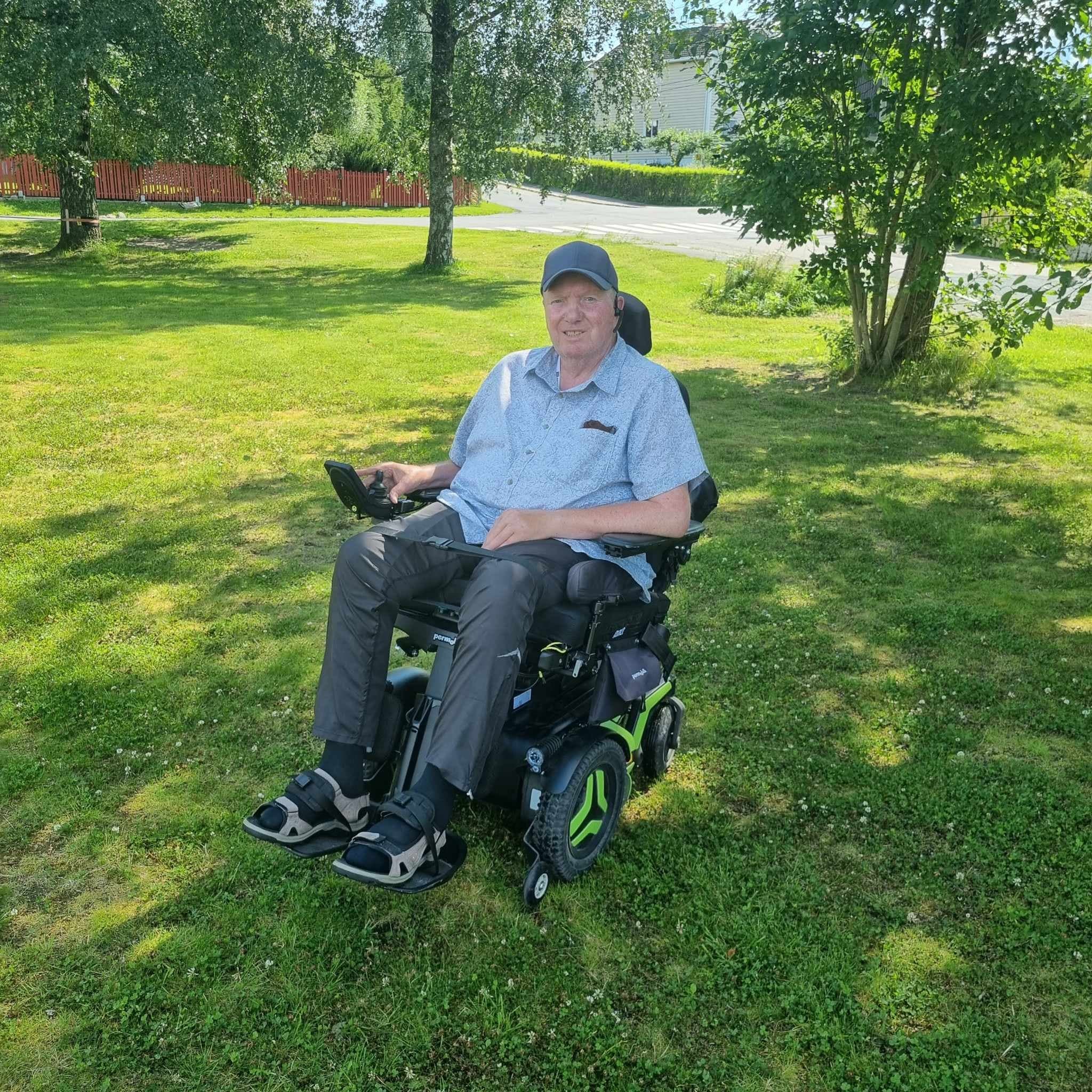 Person i motorisert rullestol med grønne hjuldetaljer i grønt parkområde med trær, gjerde og hus i bakgrunnen.